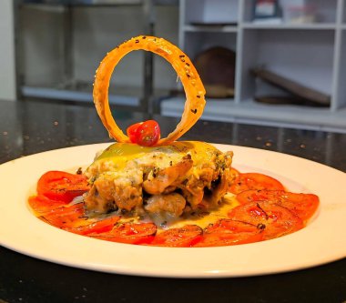 Eritilmiş peynirle kaplı istiflenmiş et ve yumurta sarısı baharatlı domates dilimleri ve beyaz bir tabakta düşey çıtır soğan halkası ile kaplıdır..