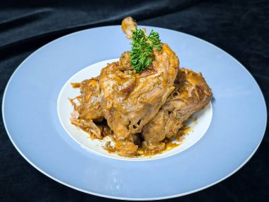 Işıl ışıl kahverengi bir sosla kaplanmış haşlanmış tavuk parçaları mavi kenarlı ve taze maydanozla süslenmiş beyaz çerçeveli tabakta servis edilir.