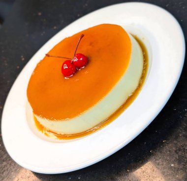 Üstü parlak karamel ve iki maraska kirazı ile kaplı klasik kremalı pasta, beyaz bir tabakta sunuluyor, temiz ve zarif bir görünüm için..