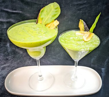 Avokado dilimi, kızarmış muz ve ıspanak yaprağıyla süslenmiş berrak bir kadehte servis edilen kalın yeşil bir smoothie..