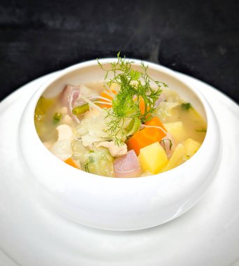 Yürekli sebze ve doğranmış tavuk çorbası havuç, patates, yeşil fasulye ve dereotlu aynı tabakta beyaz bir seramik kasede servis edilir..