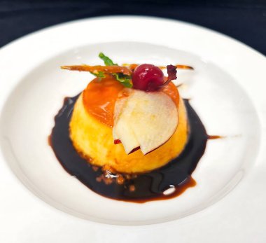 Üzerinde parlak bir kiraz, nane filizi ve karamelize şeker parçasıyla karamel turtası, koyu şerbetli beyaz bir porselen tabakta servis edilir..