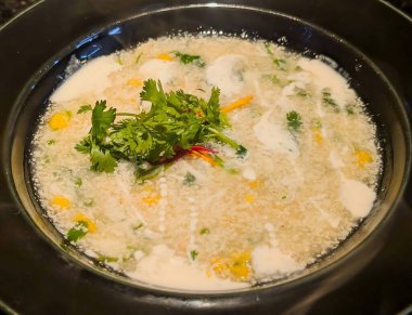 Mısır taneli kremalı mısır çorbası, doğranmış sebze ve taze kişniş, beyaz kremalı siyah bir kasede servis edilir..