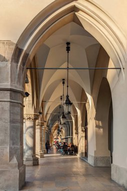 Polonya 'nın Krakow kentindeki tarihi Sukiennice atari salonunda, atlama tavanları, taş sütunlar ve asılı fenerlerle, insanlar öğleden sonra ışığında bir kafede oturuyorlar..