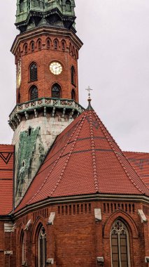 Saat yüzlü, kemerli pencereli ve bulutlu bir günde dik kiremitli çatılı kırmızı tuğlalı kilise kulesinin yakın görüntüsü..