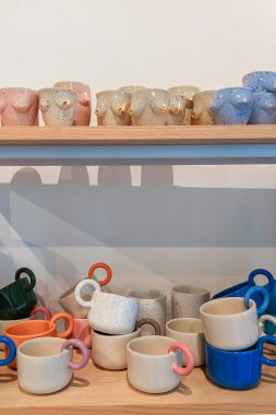 Renkli el yapımı seramik kupalar ve ahşap raflarda altın aksanlı gövde şekilli bardaklar ve zıt kulplu minimalist benekli kupalar..