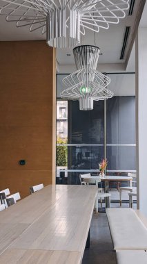 Modern yemek alanı, uzun mermer tepeli masa, beyaz sandalyeler, minimalist dekor, ve şık bir şehir kafesinde tavandan sarkan çarpıcı geometrik kolye ışıkları.