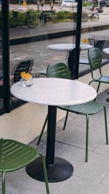 Modern yuvarlak bir terrazzo masası, yeşil metal sandalyeler, ve sarı bir çiçek taşıyan küçük cam bir vazo, şehrin kaldırımında yansımaları ile yakalandı..