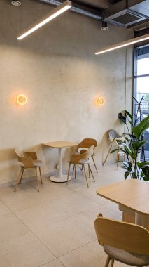 Minimalist tasarımı olan parlak, modern bir kafe. Ahşap sandalyeler ve masalar, yumuşak bej duvarlar, sıcak aydınlatma armatürleri ve geniş pencereler rahat ve şık bir atmosfer yaratır. Doğal dokuları ve kapalı bitkileri olan İskandinav esinlenilmiş bir dekor..