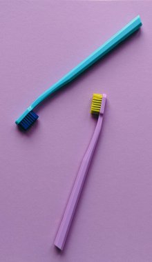 Renkli iki diş fırçasının rengarenk minimalist kompozisyonu. Bir diş fırçası koyu tüylü mavi, diğeri sarı tüylü lavanta, çapraz yerleştirilmiş..