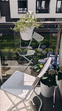 Sıcak, modern bir balkon, beyaz katlanır bir sandalye, saksı çiçekleri ve güneşli bir günde yansıyan cam korkuluklar..