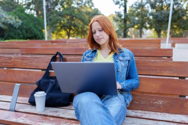 Kızıl saçlı genç kadın dizüstü bilgisayar kullanıyor, tahta bir bankta kahve ve çantayla oturuyor, teknolojiyle iç içe.