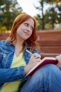 Kızıl saçlı, burnu piercing 'li, gülümseyen, açık havada deftere yazan, ilham veren genç bir kadın.