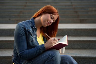 Kızıl saçlı kadın günlüğüne dalmış ya da ödevlerini yapıyor, beton basamaklarda oturuyor.