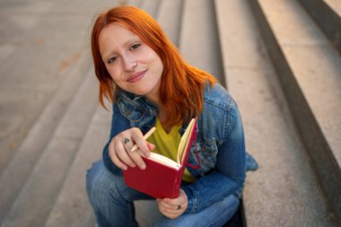 Kızıl saçlı genç bir kadın dışarıda merdivenlerde oturuyor, elinde kırmızı bir defter ve kalem var, kameraya gülümsüyor.