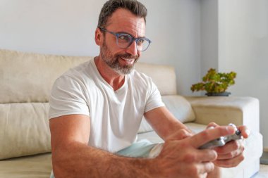 Orta yaşlı adam evdeki kanepede video oyunları oynarken gülümsüyor
