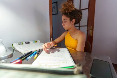 Genç öğrenci bir deftere özenle yazıyor, yaklaşan sınav için hazırlanırken evdeki masa lambasının yumuşak parıltısına odaklanıyor.