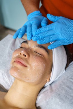 Güzellik uzmanı köpüğü mavi eldivenle temizliyor. Hastanın yüzünü profesyonel ultrasonik cilt tedavisi için hazırlıyor.