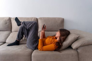 Kanepede rahat bir şekilde uzanan genç bir kadın rahat oturma odasında rahat ve mutlu bir hava yayarken akıllı telefonunun tadını çıkarıyor.