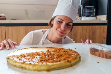 Aşçı şapkası ve önlüğü takan bir kadın, modern bir mutfakta tarçınlı büyük yuvarlak bir pastayı zevkle izliyor.