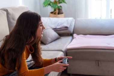 Gülümseyen bayan oyuncu video oyunundan zevk alıyor. Oturma odasının zemininde rahatça otururken kontrolörü sıkıca kavrıyor.