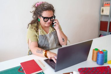 Terzisi dikiş atölyesinde çalışıyor. Dizüstü bilgisayarındaki siparişleri yönetiyor ve telefonda müşterisiyle konuşuyor.