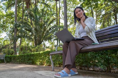 Genç Japon kadın parktaki bankta otururken dizüstü bilgisayar kullanıyor, temiz havanın ve yeşil çevrenin tadını çıkarıyor.