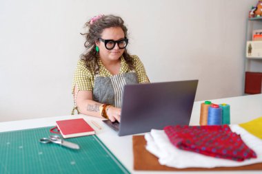 Rahat bir dikiş atölyesinde dizüstü bilgisayarda çalışan, çevrimiçi siparişleri yöneten ve küçük işletmesi için sosyal medyayla uğraşan bir terzi.