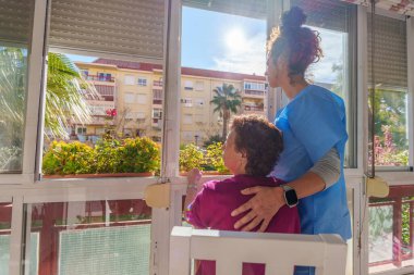 Destekleyici bakıcı yaşlı kadına huzur sunuyor, pencereden bakıyor, gündüz saatlerinde sıcaklığı ve şefkatli bakımı paylaşıyor.