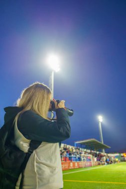 Orta yaşlı bir kadın spor fotoğrafçısının Alacakaranlık 'ta stadyum ışıkları ve gökyüzü ile futbol sahasında fotoğraf çekerken görüntüsü