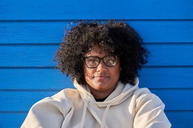 Güneşli bir günde mavi arka planda bereli kazak ve afro saç giyen siyahi bir kadın. 