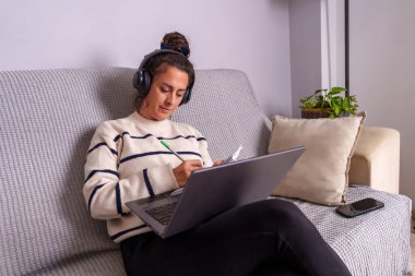 Kulaklık takan bir kadın çevrimiçi kursa katılırken not alıyor. Laptop kullanıyor, rahat oturma odasındaki kanepede çalışıyor.