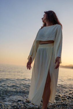 Gün batımında çakıl taşlı kıyı şeridinde duran kadın, altın ışığa karşı silueti olan, kıyı dinginliğini ve okyanusun huzurlu manzarasını deneyimleyen kadın.