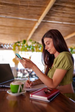 Freelancer not alıyor ve bilgisayarlı ve cappuccinolu bir barda çalışırken akıllı telefon kullanıyor.