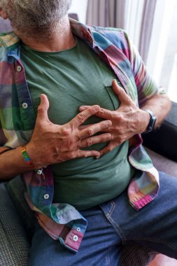 Yaşlı erkek göğsünü sıkıyor, oturma odasındaki kanepede dinlenirken ciddi kalp rahatsızlığından yüzünü buruşturuyor.