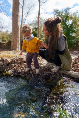 Genç bir anne, oğlunun ormanda doğayı keşfetmesine yardım ediyor, güneşli bir günün tadını çıkarıyor.