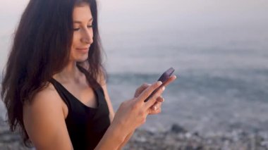 Spor kıyafetli kadın, altın gün batımında kumlu sahilde okyanus dalgaları yakınında huzurlu bir anın tadını çıkarırken akıllı telefonuna göz atıyor.