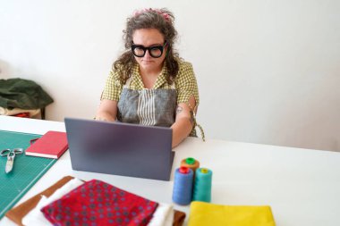 Ev atölyesinde canlı iplik makaraları ve desenli kumaş örnekleriyle çevrili dizüstü bilgisayarda çalışan kadın terzi.