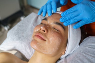 Güzellik uzmanı genç kadının yüzüne temizleyici krem sürüp ultrasonik yüz tedavisine hazırlıyor.