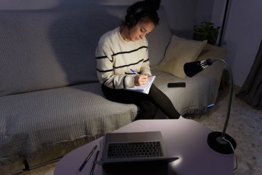 Geç saatlere kadar çalışan, kulaklık takan, dizüstü bilgisayardan ders notları alan ve akşam saatlerinde kanepede dinlenen bir kız öğrenci.