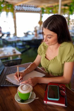 Modern bir barda bilgisayarla uzaktan çalışıp cappuccino içen serbest çalışan kadın notlar yazıyor.