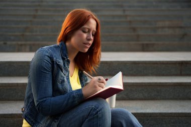 Konsantre olan genç bir kadın, bir günlükle notlar yazarken beton merdivenlerde oturuyor, dışarıda öğreniyor.