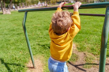 Parlak sarı kapüşonlu kapüşonlu bir çocuk yatay barı tutuyor, dışarıda oyun oynarken şakacı bir şekilde sallanıyor.