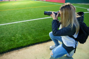 Bir futbol sahasında Alacakaranlık Maçı sırasında fotoğraf çekmek için çömelmiş orta yaşlı bir kadın spor fotoğrafçısının yan görüntüsü