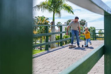 Babası, şehir parkında oğluyla motosiklet sürerken, güneşli bir günün tadını çıkarırken gülüp kaynaşıyor.