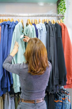 Bir mağazada kıyafet arayan bir kadının arka görüntüsü, açık yeşil kapüşonlu bir askıya dokunuyor ve bir sürü renkli kazak giyiyor.