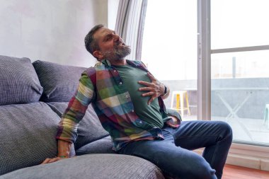 Sakallı bir adam rahatsızlık yaşıyor ve göğsünü sıkıyor. Potansiyel bir kalp krizi ya da başka bir acil durum belirtisi.