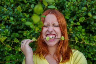 Kızıl saçlı genç bir kadın, yüzünü buruşturuyor, yeşil bir bitki dalını ısırıyor. Doğal ortam, çaba gösterme ve doğayla eğlenceli etkileşim