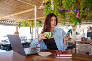 Genç yönetici kahve içiyor ve restoranda laptopuyla çalışırken akıllı telefon kullanıyor.