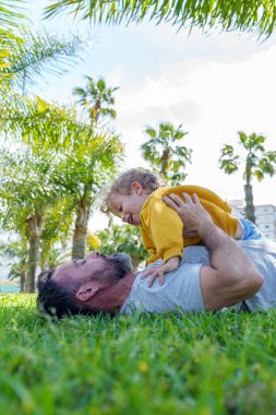 Baba çimenlerin üzerinde uzanmış güneşli bir parkta oynarken oğlunu neşeyle kucağında tutuyordu. Palmiye ağaçları ve yeşilliklerle çevriliydi.